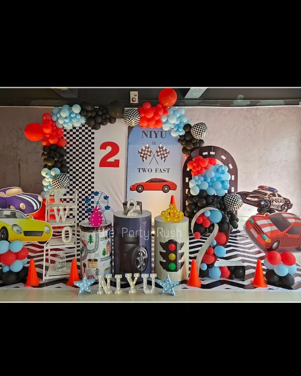 This 'Two Fast' racing theme for Niyaan is ready for the fast lane. The setup includes a checkered flag backdrop, multiple car cutouts, a custom-designed floor mat that looks like a zebra crossing, and a balloon arch that mimics a finish line.