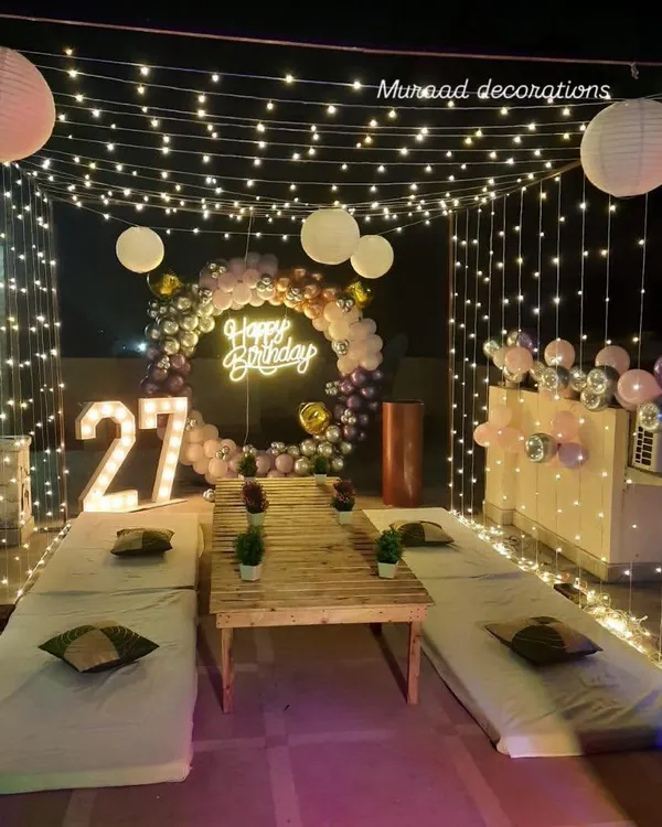 This terrace was converted into a stylish party lounge for a 27th birthday. The setup includes a fairy light canopy, low seating, a balloon ring with a neon sign, and large marquee numbers.