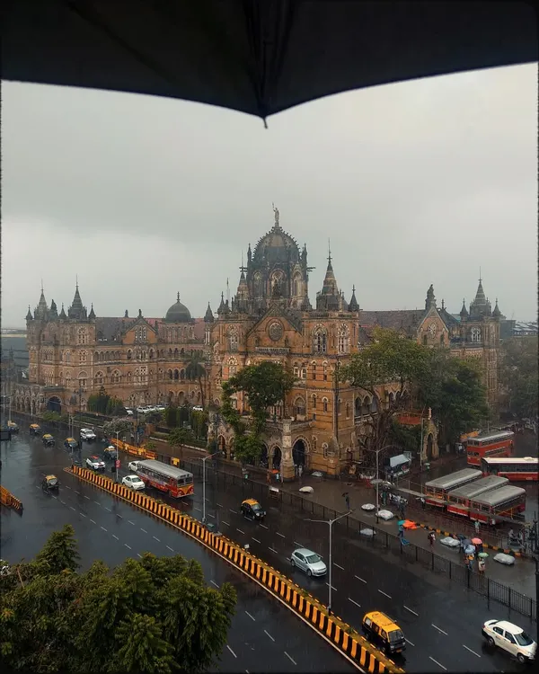 A classic monsoon view of the Chhatrapati Shivaji Maharaj Terminus, framed by the top of an umbrella. This perspective makes the viewer feel like they are right there, witnessing the grandeur of the architecture through the rain.