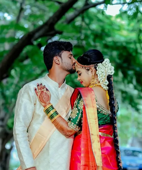 A tender forehead kiss shared between a couple in beautiful traditional attire. The bride's red and green silk saree and the groom's cream sherwani create a classic and elegant look.