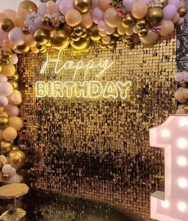 A glamorous first birthday backdrop featuring a wall of gold sequins. The setup is enhanced with a 'Happy Birthday' neon sign, a light-up marquee number '1', and a beautiful balloon arch in gold and pastel pink.