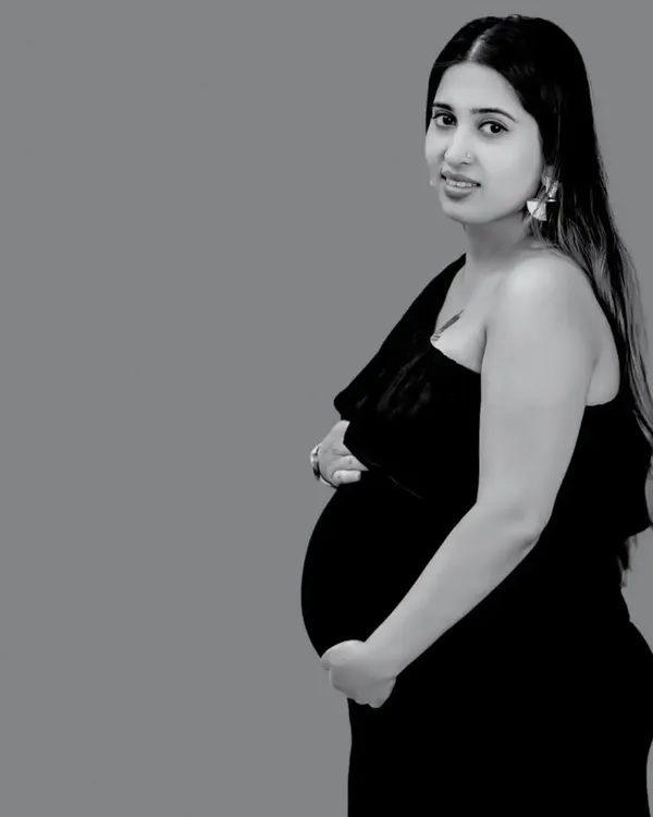 A classic black and white solo portrait. The simple, elegant pose and one-shoulder dress create a timeless silhouette, focusing all attention on the mother's gentle expression and her beautiful baby bump.