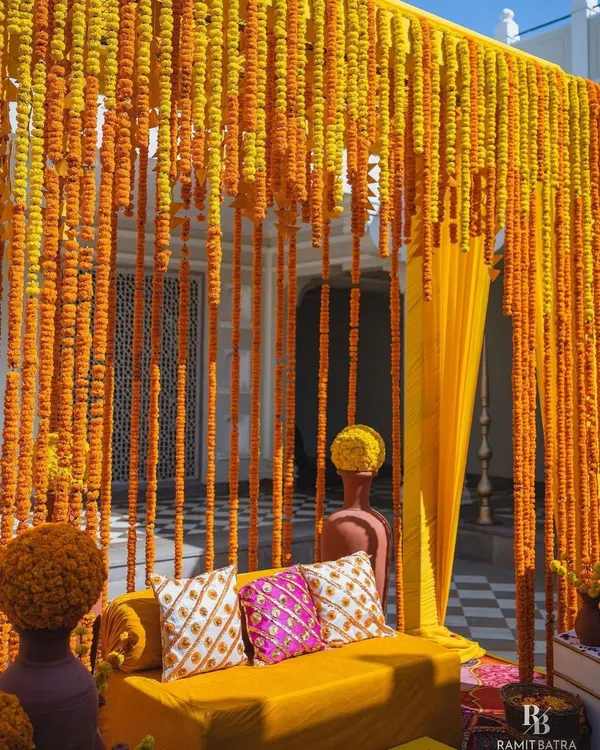 A classic Haldi seating area, defined by a dense curtain of fresh marigold strings. The vibrant yellow sofa and patterned cushions complete this traditional and picture-perfect photo corner.