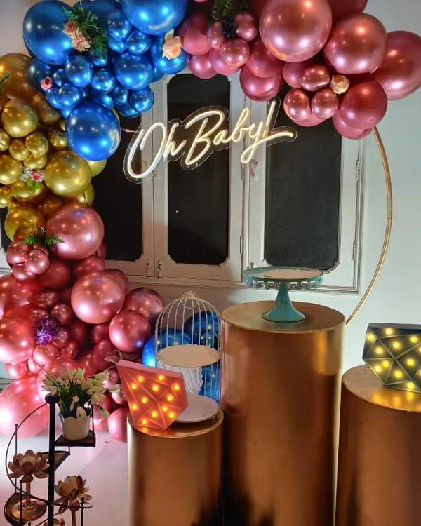 A glamorous 'Oh Baby' shower setup with a circular ring, a cascade of chrome balloons in gold, blue, and pink, and elegant copper-toned plinths.