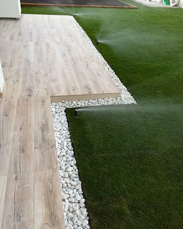 A modern backyard featuring a flawless lawn with an integrated pop-up sprinkler system in action. The lawn is edged with white pebbles, creating a sharp, clean border against the wood-look tile deck.