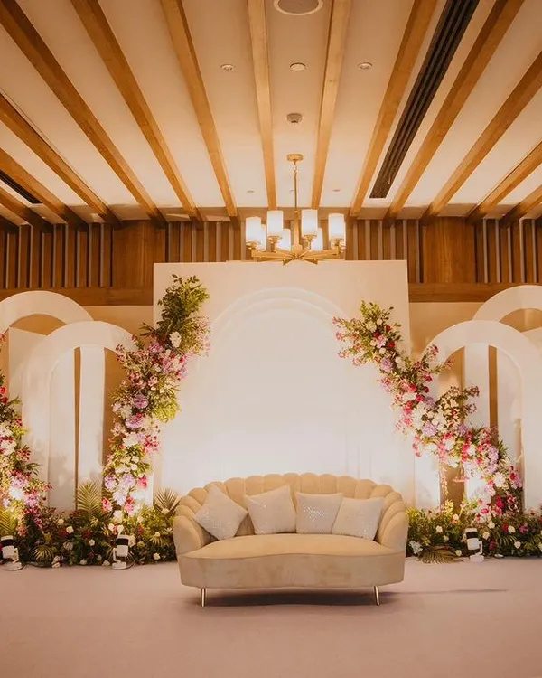 A beautiful and elegant stage for an engagement or Roka ceremony, featuring white arches, blush floral arrangements, and a classic scalloped sofa. The design is soft, romantic, and sophisticated.