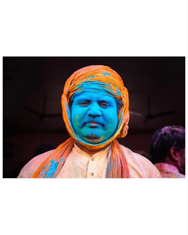 A man's face, covered in blue Holi powder, offers a moment of calm contemplation amidst the festival's chaos. His orange turban provides a striking color contrast.