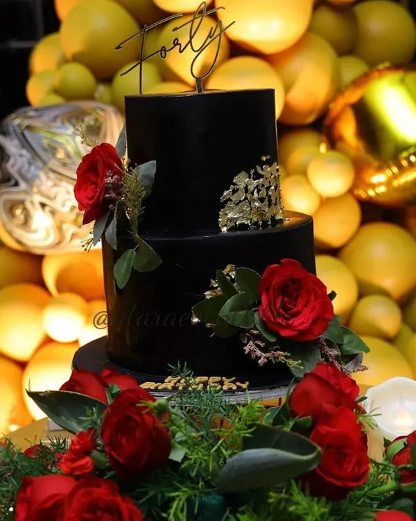 A stunning two-tiered black cake for a 40th birthday celebration. The cake is elegantly decorated with deep red roses and flecks of gold leaf, topped with a "Forty" sign, making it a sophisticated centerpiece.