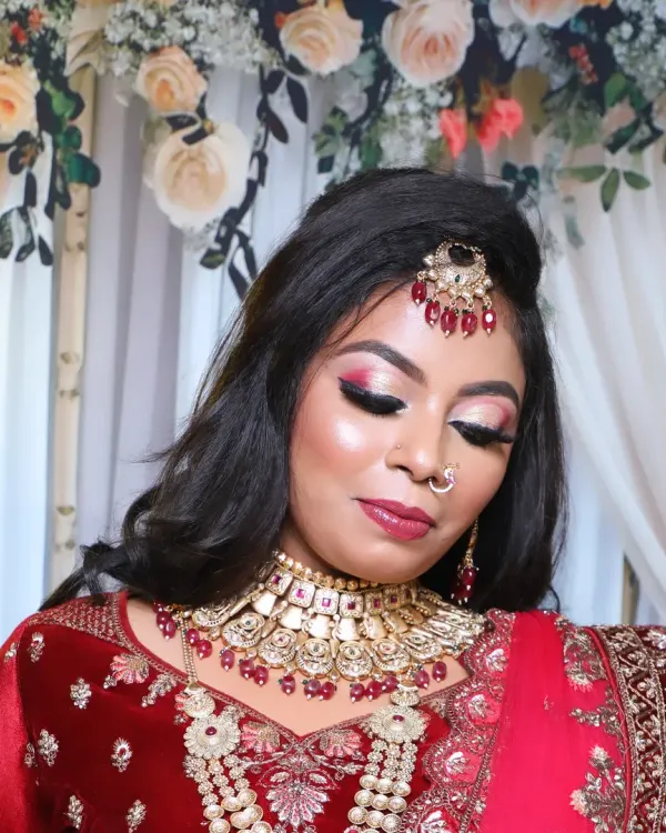 A close-up of a glamorous bridal look in a red lehenga. The makeup features dramatic red and pink eyeshadow, sharp contouring, and a full set of jewellery including a maang tikka and choker.