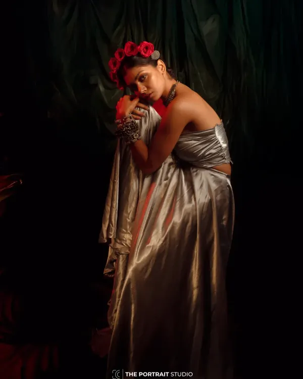 A portrait from the 'INARA' series, styled to evoke a sense of romantic melancholy. The combination of a liquid silver top, heavy traditional jewelry, and a crown of red roses creates a powerful, goddess-like image.