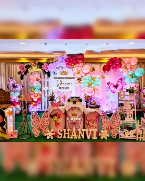 A beautiful and colorful butterfly theme for Shanvi's first birthday. The setup includes a lush green carpet, large butterfly wing cutout's, fairy character's, and a vibrant balloon backdrop.