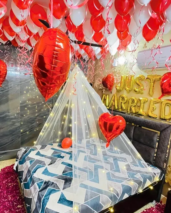 A classic romantic room setup for newlyweds. The ceiling is covered with red and white balloons, while a delicate fairy light canopy drapes over the bed. 'Just Married' foil balloons and heart-shaped balloons complete the look.