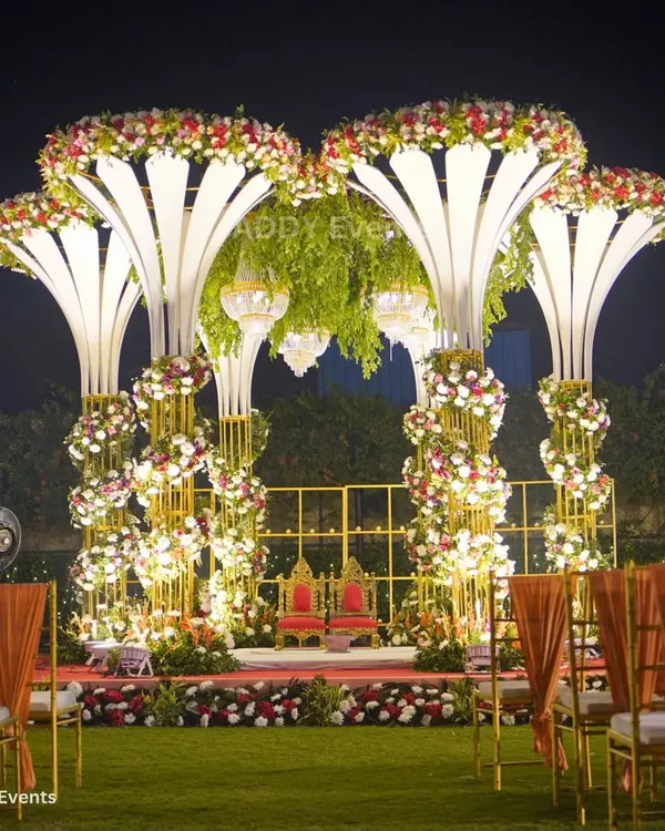 A mandap that commands attention. This magnificent structure features illuminated floral-inspired pillars, a canopy of lush greenery, and suspended crystal chandeliers, creating a royal setting for an unforgettable night wedding.