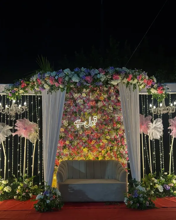 A breathtaking Nikah stage at night. The backdrop is a dense wall of colorful flowers with a 'Qubool Hai' neon sign, framed by a floral arch, white drapes, and sparkling string lights.