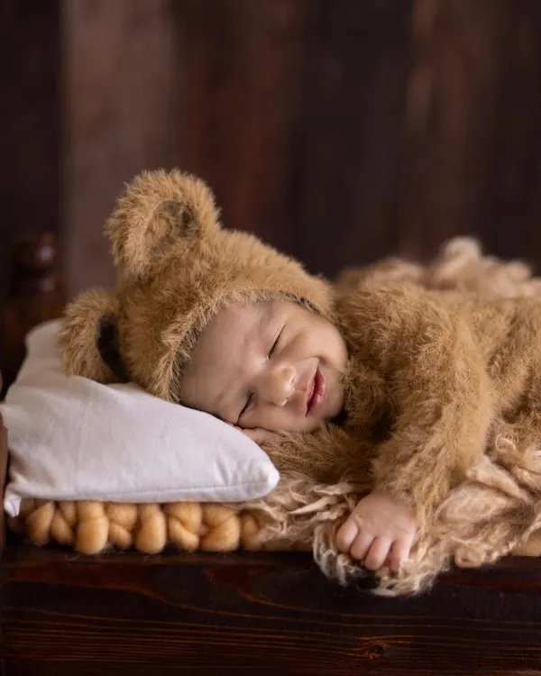 A heartwarming close-up of a smiling newborn dressed in a fuzzy teddy bear outfit, sleeping soundly on a tiny pillow. This shot captures the pure, innocent joy of a sleeping baby.