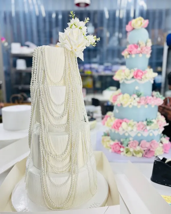 A beautiful comparison of two of our grand celebration cakes. On the left, a four-tier white cake with elegant pearl draping, and on the right, a five-tier pastel blue cake adorned with hundreds of handcrafted sugar flowers.