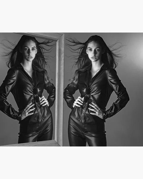 A conceptual black and white fashion portrait featuring a model in a leather outfit. The use of a mirror and wind creates a dynamic, high-energy image suitable for an edgy fashion editorial.
