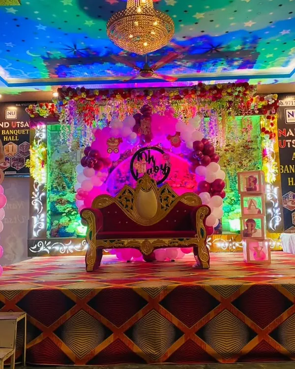 A grand stage setup for a naming ceremony in a banquet hall. The decor includes a royal-style sofa, Boss Baby character cutouts, and pink and white balloon pillars, creating a special spot for the main event.