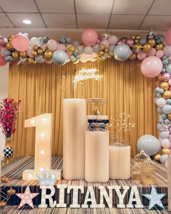 We created this beautiful setup for Ritanya's first birthday party. It features a custom balloon arch in pastel pink, blue, and gold, a glowing 'Happy Birthday' neon sign, and a large light-up number '1'. The little white picket fence and decorative plinths for the cake and props added a charming touch to the entire photo corner.