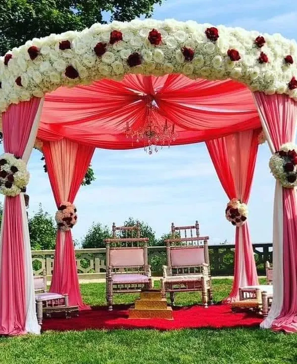 An elegant outdoor Vidhi Mandap we designed at a resort, featuring a beautiful floral canopy of white and red roses with flowing pink drapes. We create sacred and beautiful spaces for you to take your vows.