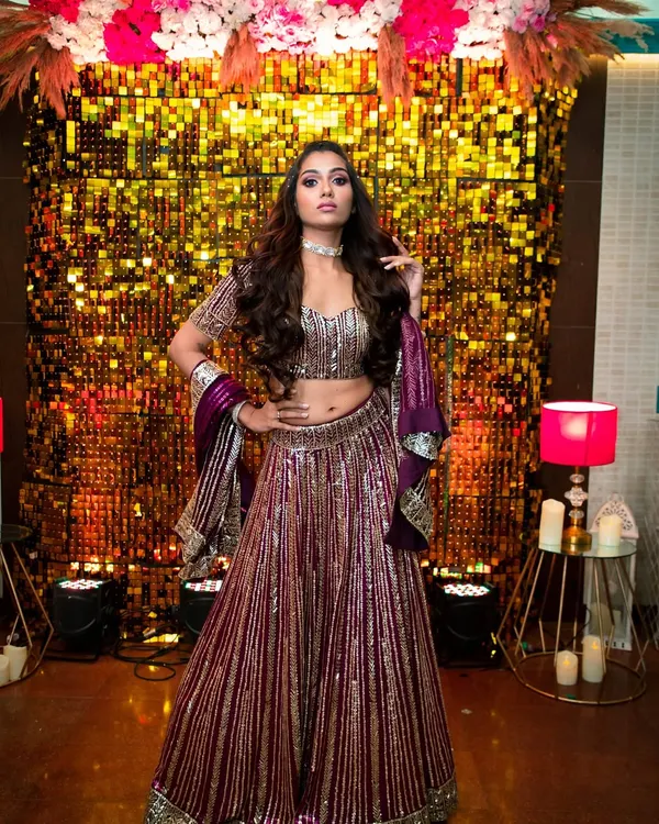 This is a full-on glamour look perfect for a sangeet or reception night. The makeup features a dramatic, colorful smokey eye and contoured cheeks, set against a glittering backdrop that matches her energy.