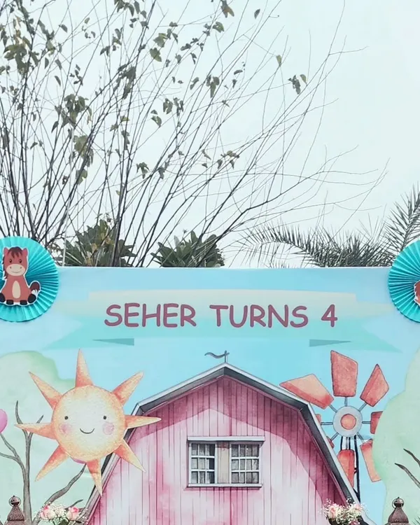 A close-up of the farm-themed birthday backdrop for "Seher Turns 4." The design features a cheerful sun, a pink barn, and a windmill, all in a cute, storybook art style.