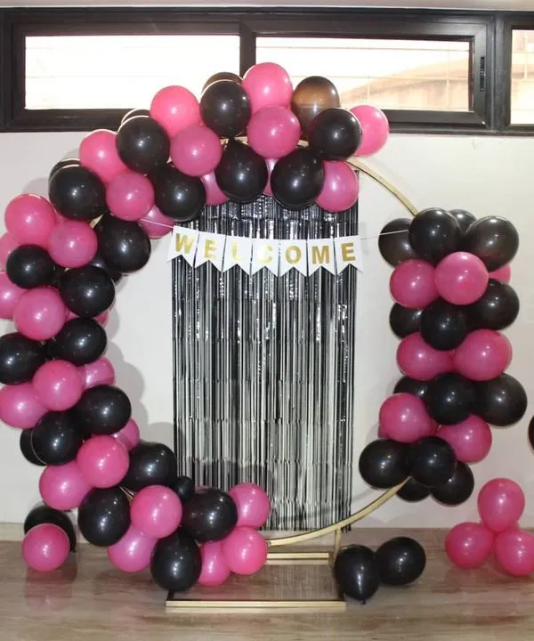 A fun and modern welcome home decoration. This setup features a circular frame with a pink and black balloon arch, a 'Welcome' banner, and a silver fringe curtain for a touch of sparkle.