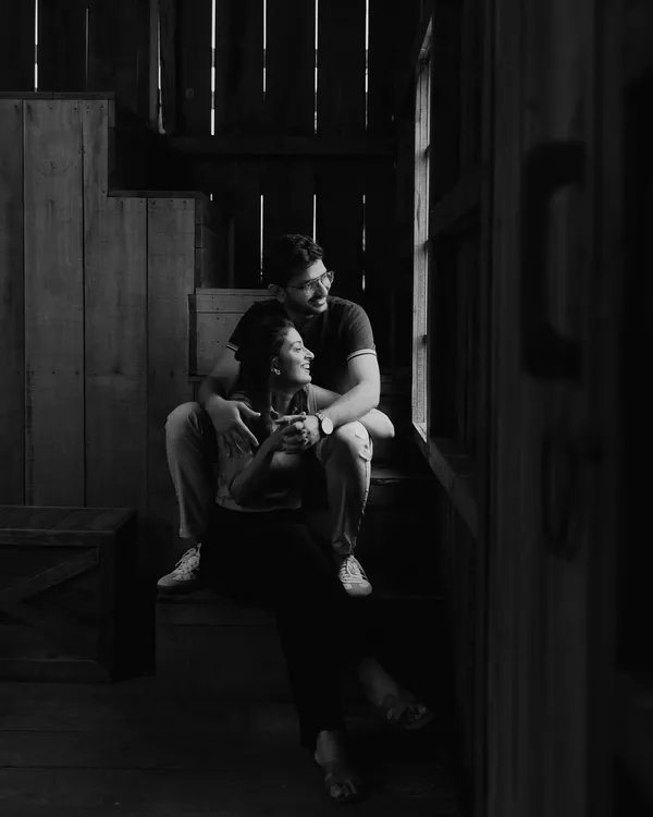 A quiet, contemplative moment captured in black and white. The couple sits by a window, bathed in soft, natural light, creating a timeless and soulful portrait.