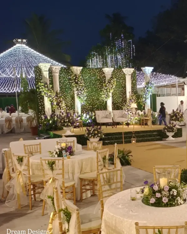 An evening reception at the Colaba Agiary. The space is illuminated by a canopy of fairy lights, with a grand stage featuring a green wall and classical pillars. The guest tables are elegantly set to complete the luxurious atmosphere.