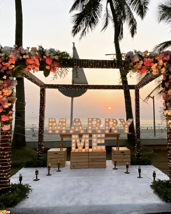 The perfect proposal setting at sunset. A beautiful floral arch frames the sea view, with 'MARRY ME' marquee lights ready for the big question.