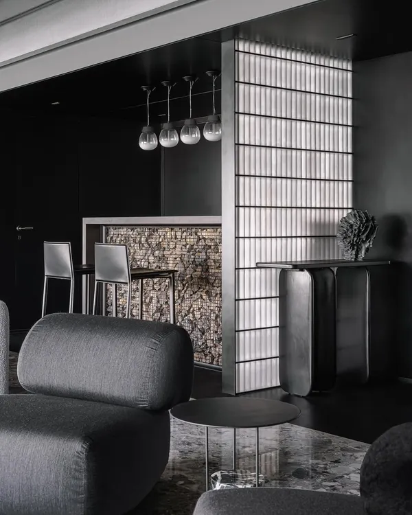 A full view of the bar in the Vaulted Haus, showing the Gabion wall facia, frosted glass brick partition, and custom pendant lights. The entire unit is conceived as part of a black 'scoop' that carves through the apartment's entrance.