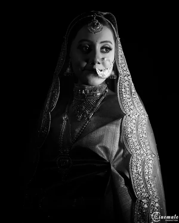 A dramatic black and white bridal portrait using high-contrast lighting to create a powerful and mysterious image. The focus is entirely on her expression and the intricate details of her attire.