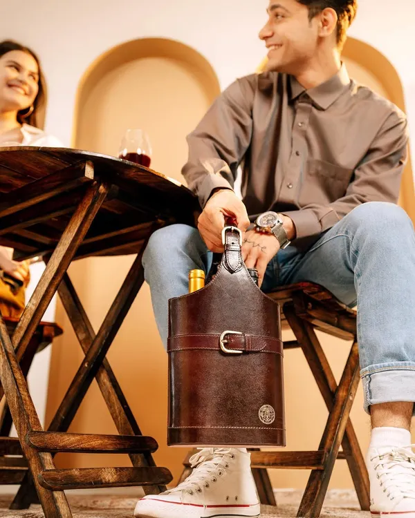 A lifestyle scene for Verman Atelier's leather wine carrier. The shot captures a candid moment between a couple, with the product looking stylish and functional in a social, sophisticated setting.