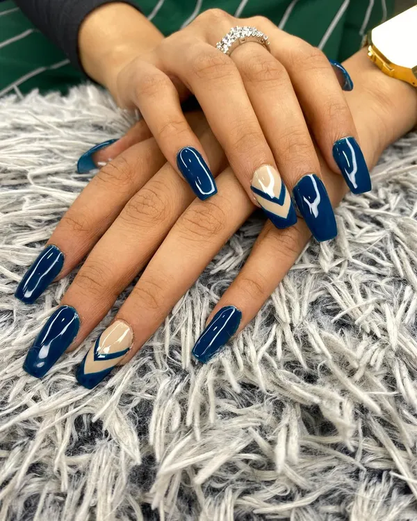 This manicure features a bold navy blue gel polish on long, coffin-shaped nails. The accent nails use a negative space technique with a nude base and sharp, geometric blue chevrons for a modern edge.
