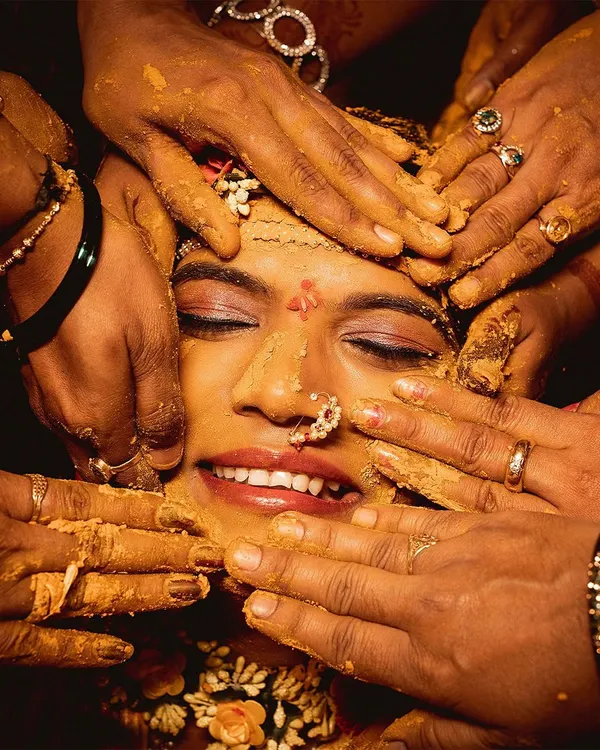A close up of the bride during her Haldi ceremony. Surrounded by the hands of her loved ones applying turmeric paste, her smile shows pure happiness.