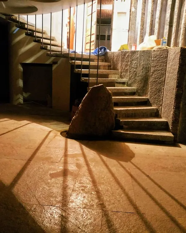 The saga of stones and shadows. In this basement parking area, simple lighting casts dramatic shadows from the railing, turning a functional space into a theatrical one. A natural boulder anchors the stone staircase, adding an element of raw nature.