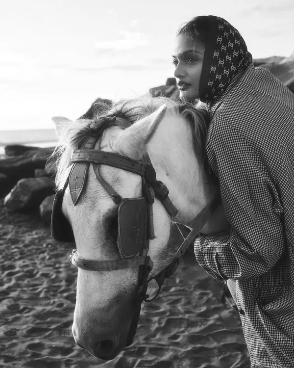 A black and white editorial fashion shot featuring a model in a headscarf and houndstooth jacket, sharing an intimate moment with a white horse on a sandy beach. This image captures a timeless, cinematic feel.