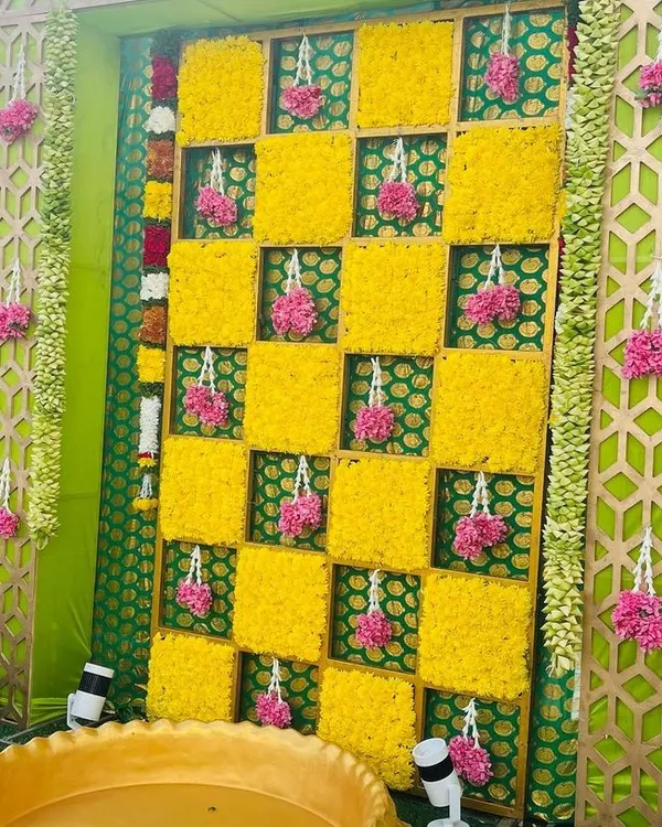 A vibrant Haldi backdrop with a checkerboard pattern of yellow marigolds. The setup includes a traditional brass vessel (urli) for the ceremony, framed by decorative panels.