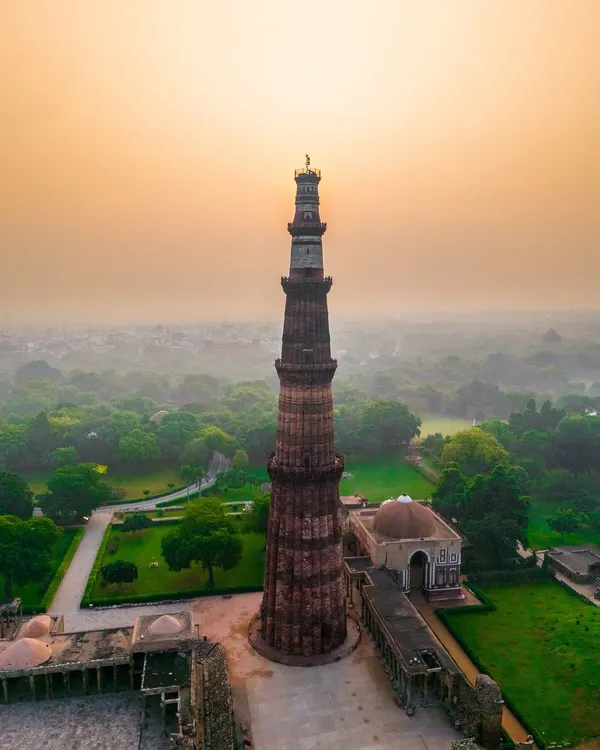 The Qutub Minar in Delhi stands tall against a hazy sunrise. This aerial shot captures the monument and its surrounding lush greenery from a unique, elevated perspective.