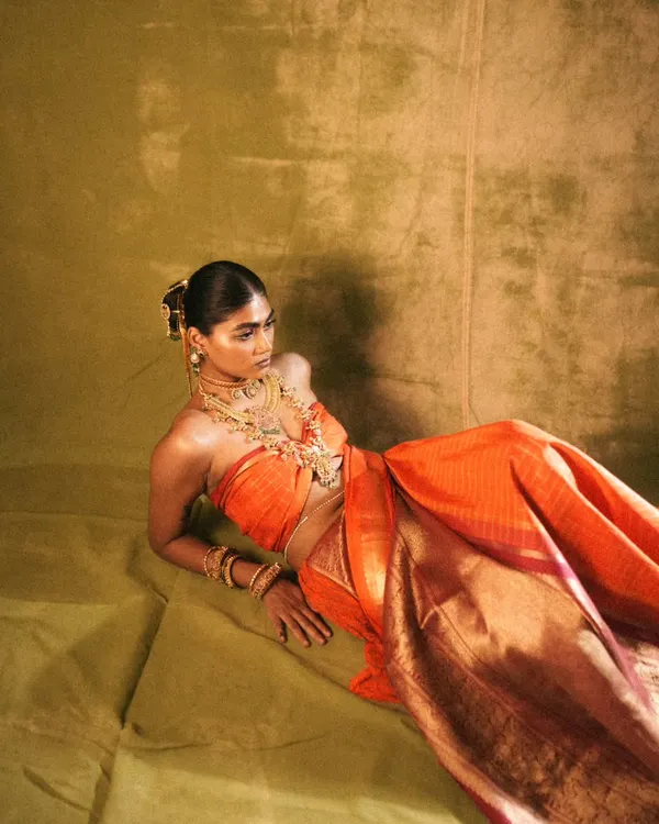 A full-length view of the 'Swaṟṇavalli' concept, where the rich orange of the silk sari is complemented by warm, bronzed skin and traditional gold jewellery. The makeup was designed to feel both regal and ethereal.
