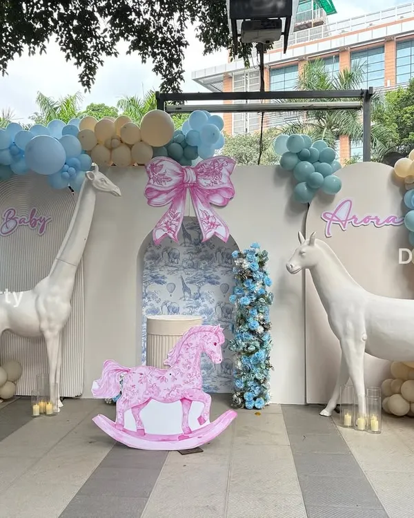 A grand Dior-inspired baby shower backdrop with a soft blue and pink color scheme. The setup features life-sized animal props like a giraffe and unicorn, a rocking horse, and a vintage-style pram cutout for a touch of elegance.