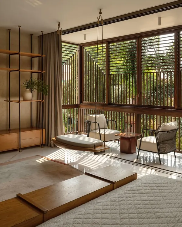 This bedroom is designed as a tranquil retreat, where the morning sun filters through timber lattices, creating a dynamic play of light and shadow. The space opens onto a modern 'jharokha' or enclosed balcony, featuring a traditional swing, blurring the line between the comfort of the indoors and the serenity of the outdoors.