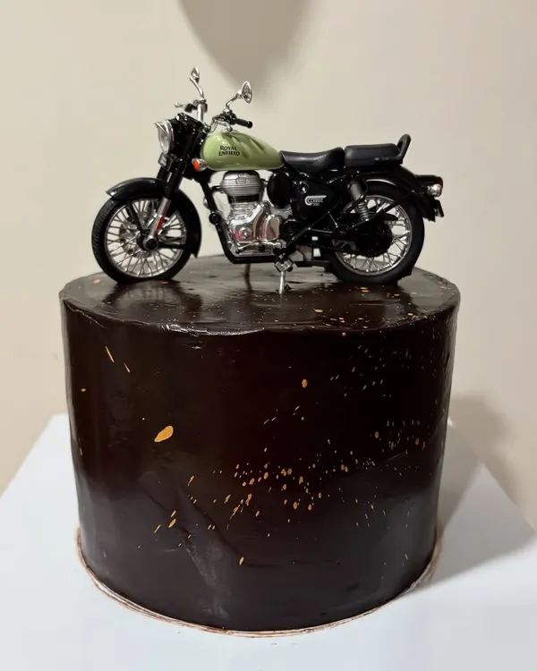 A tall 1.5kg Chocolate Ganache cake for a bike lover, topped with a detailed Royal Enfield model. The dark chocolate glaze with gold splatter gives it a sleek, masculine look.
