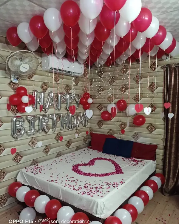 A classic romantic birthday room decoration with red and white balloons on the ceiling and floor, "Happy Birthday" foil letters, and a heart made of rose petals on the bed.