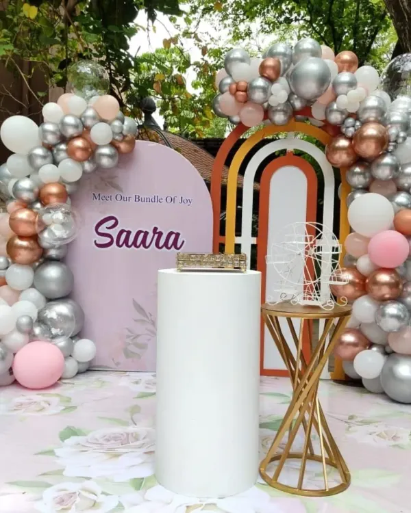 A close-up of a modern and chic naming ceremony backdrop for "Saara". The decor features a pastel color palette with rose gold, silver, and pink balloons, a custom arched backdrop, and elegant pedestals.
