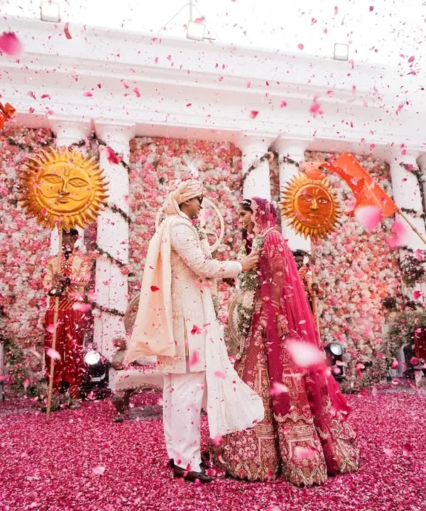 A perfect Varmala moment captured amidst a shower of pink flower petals. The couple is framed by dancers with sun props and a stunning floral wall, creating a scene of pure romance and grandeur.