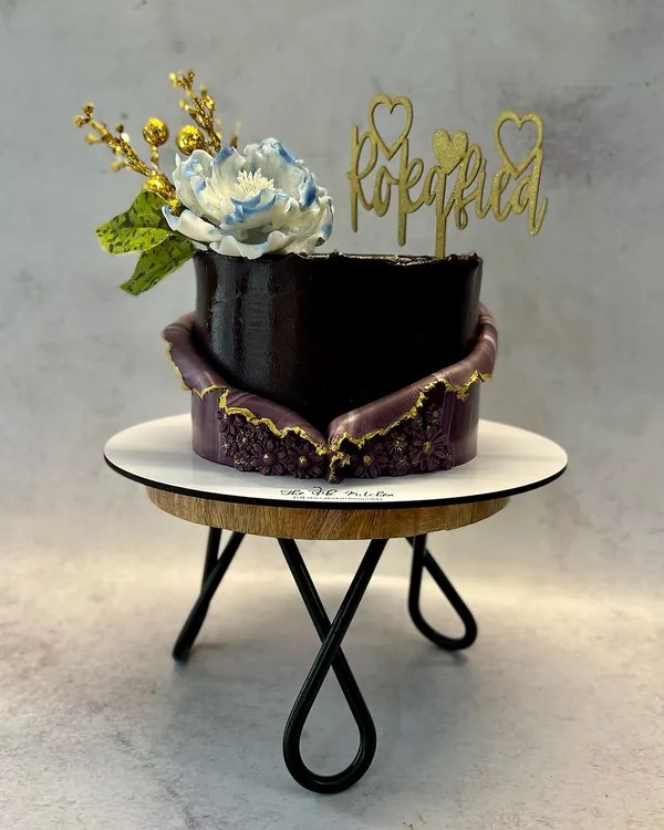 This modern Roka cake features a rich, dark chocolate finish with a stunning purple geode-style fault line, edged in gold. The design is finished with a delicate sugar flower and a fun 'Rokafied' acrylic topper.