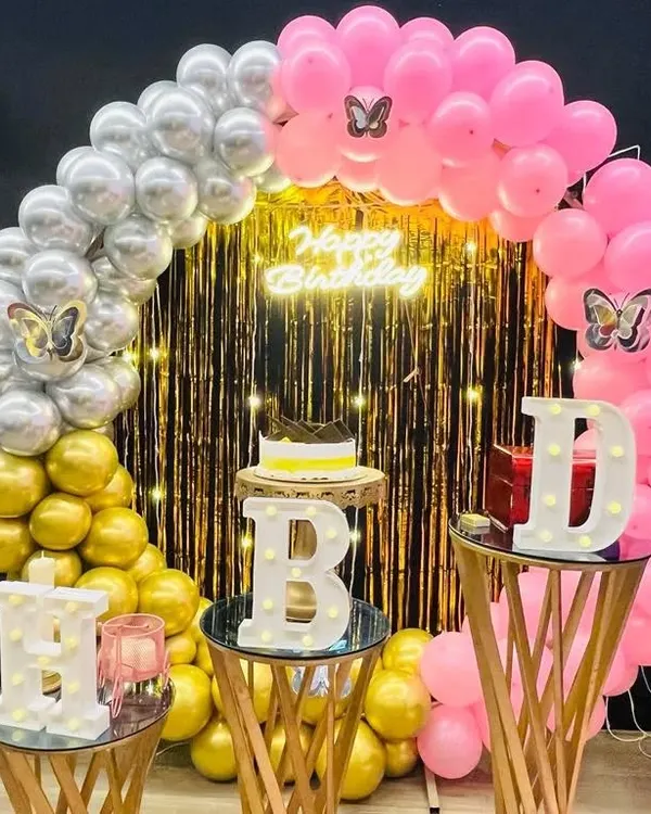 A vibrant and colorful birthday decoration setup. This image showcases a beautiful balloon arch with pink, silver, and gold, a shimmering backdrop, and our popular light-up 'HBD' letters.
