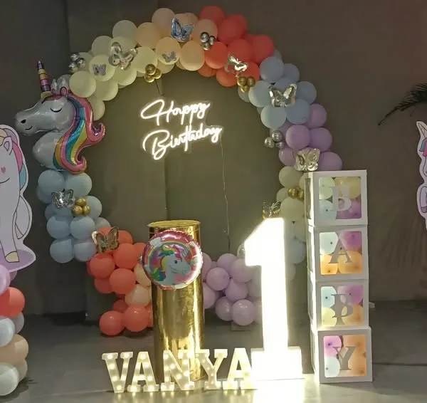 A magical unicorn-themed setup for Vanya's first birthday. It features a circular backdrop with a neon sign, unicorn cutouts, a light-up number '1', and a pastel rainbow balloon arch.