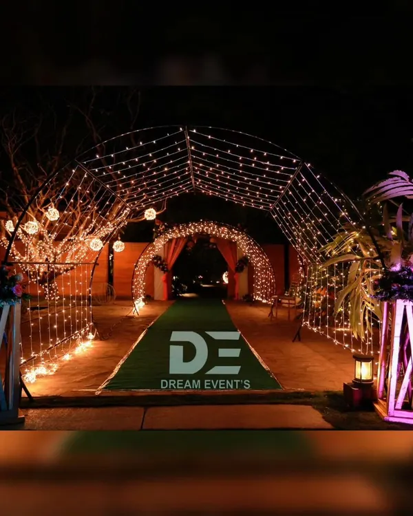 A magical entrance tunnel created with arches of fairy lights, leading guests into a wedding event over a green carpet walkway.